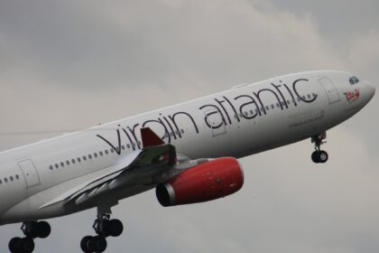 The front section of a Virgin Atlantic plane is ascending, nose to the upper right in flight. The aircraft has the airline's branding across the white fuselage, as well as red engines, and is pictured against a cloudy grey sky in the background.