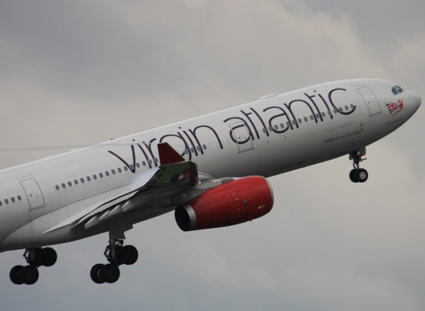 The front section of a Virgin Atlantic plane is ascending, nose to the upper right in flight. The aircraft has the airline's branding across the white fuselage, as well as red engines, and is pictured against a cloudy grey sky in the background.