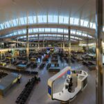The inside of terminal 2 at London Heathrow Airport, taken from above with what looks like a 0.5 lens. The top of the image shows the arched roof of the structure and its widows. below, people can be seen standing, walking, and generally congregating in the central waiting seating area, which occupies the bottom two thirds of the image. Four large pillars can be seen, two in the foreground and two in the background.