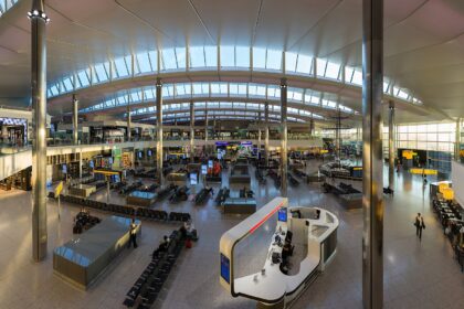 The inside of terminal 2 at London Heathrow Airport, taken from above with what looks like a 0.5 lens. The top of the image shows the arched roof of the structure and its widows. below, people can be seen standing, walking, and generally congregating in the central waiting seating area, which occupies the bottom two thirds of the image. Four large pillars can be seen, two in the foreground and two in the background.