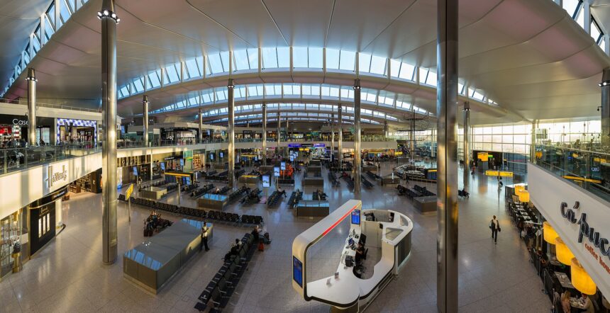 The inside of terminal 2 at London Heathrow Airport, taken from above with what looks like a 0.5 lens. The top of the image shows the arched roof of the structure and its widows. below, people can be seen standing, walking, and generally congregating in the central waiting seating area, which occupies the bottom two thirds of the image. Four large pillars can be seen, two in the foreground and two in the background.