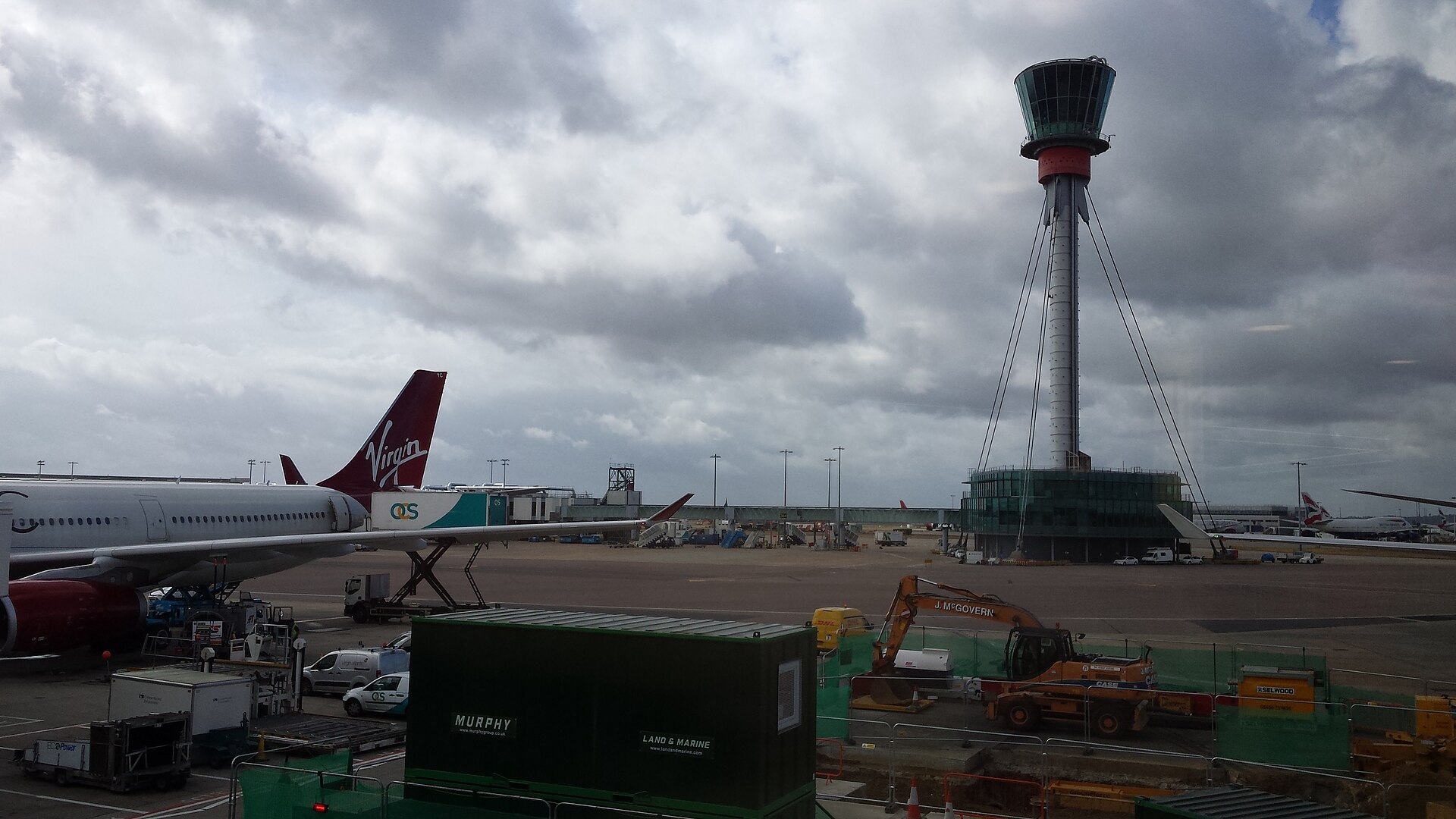 London Heathrow's airport apron is pictured. The watch tower can be seen in the centre left of the photo, and the back end of a Virgin Atlantic aircraft is visible from the far left into the centre. In the foreground, construction apparatus is fenced off to the bottom right, and a grey metal storage container is in the centre. The background depicts more of the airport apron in the distance, with grey sky occupying the majority of the background behind the watch tower. 