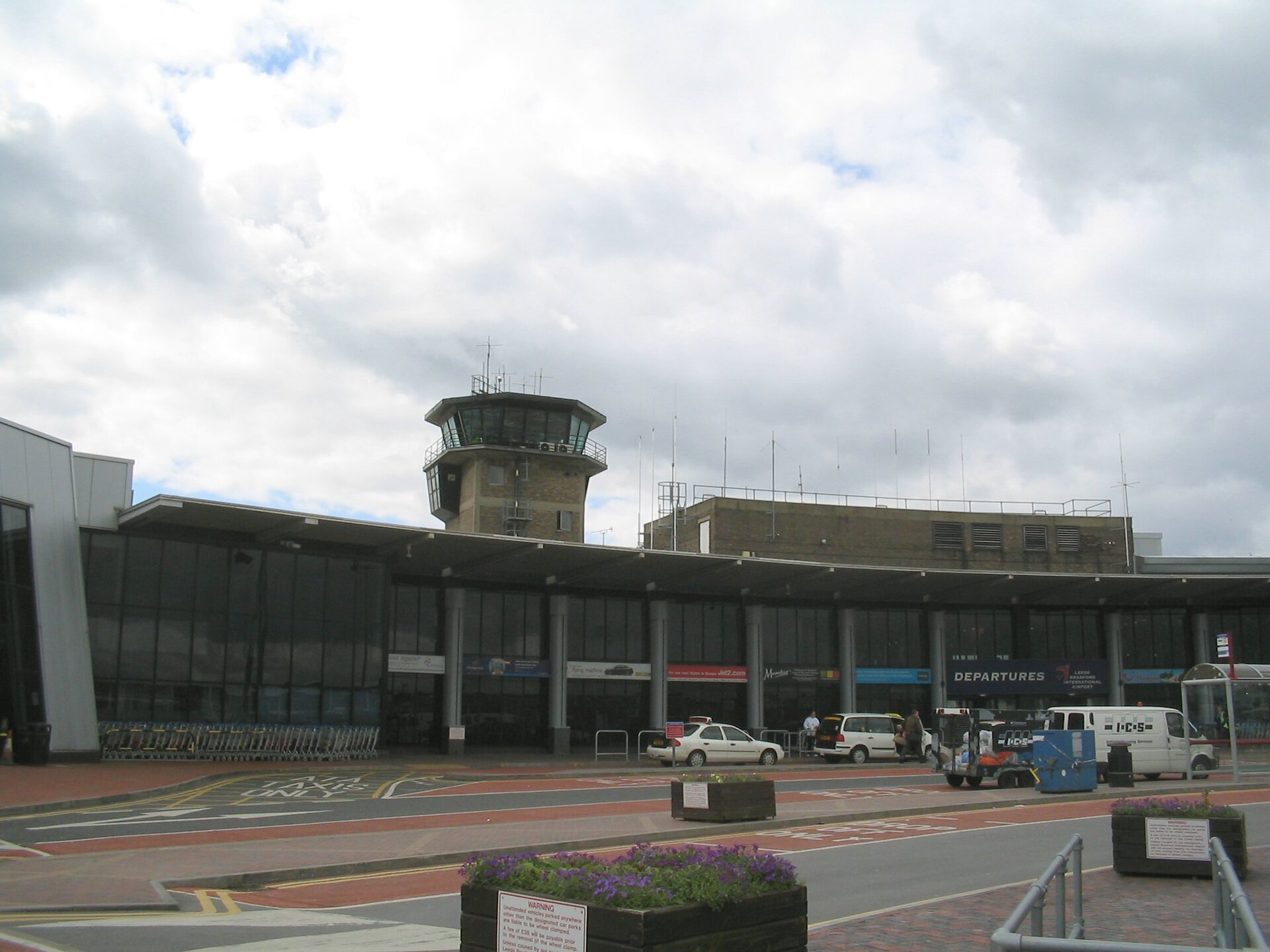 The terminal and control tower at Leeds Bradford International Airport (LBA). From the bottom of the image to the centre a road leading up to the airport building in front of the entrance can be seen with cars and vans parked up outside. The building itself is composed of glass facade walls and grey metal pillars leading up to a grey metal roof. The brick tower peeks above the building from behind in a cylindrical octagonal shape, with windows around the perimeter of the top. Another brick building stands beside the tower to the right. The upper half of the image depicts a cloudy sky.
