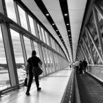 A black and white/greyscale image of people walking - taken from behind - along a moving walkway at Gatwick Airport. the walkway is on the right of the image. On the left is bright floor to ceiling windows.