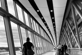 A black and white/greyscale image of people walking - taken from behind - along a moving walkway at Gatwick Airport. the walkway is on the right of the image. On the left is bright floor to ceiling windows.