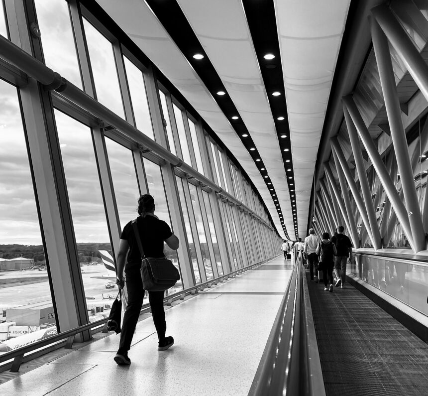 A black and white/greyscale image of people walking - taken from behind - along a moving walkway at Gatwick Airport. the walkway is on the right of the image. On the left is bright floor to ceiling windows.