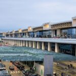 Milan Malpensa Airport pictured from he outside at either sunset or sunrise. The image is taken at an angle from the left, meaning the length of the airport can be seen large anc lose up on the right side, gradually getting smaller as it transitioned to the left size, which shows the furthest point away. The airport is lit with warm yellow lighting. the background is a bluish purple sky.