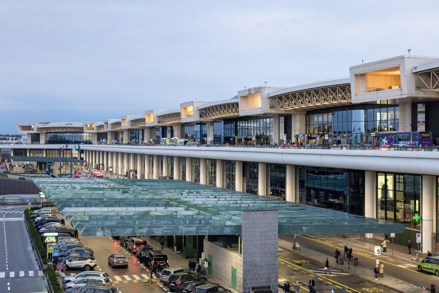 Milan Malpensa Airport pictured from he outside at either sunset or sunrise. The image is taken at an angle from the left, meaning the length of the airport can be seen large anc lose up on the right side, gradually getting smaller as it transitioned to the left size, which shows the furthest point away. The airport is lit with warm yellow lighting. the background is a bluish purple sky.