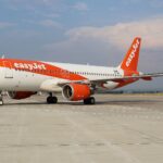 An easyJet aircraft is pictured grounded at an airport. The aircraft is mostly white, with easyJet's signature orange colouring and branding across the fuselage and tail. The aircrafts is pictured from the side closer to the front than the back at an angle, and the nose is facing left and the tail is to the right. The aircraft is positioned mostly central, with tarmac ground in he foreground and a clear blue sky with minimal cloud in the background.