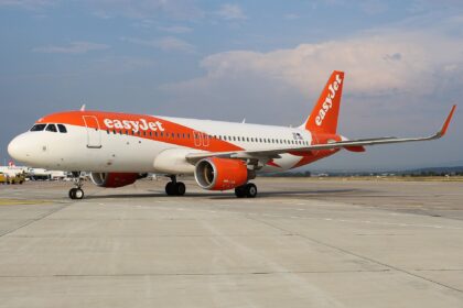 An easyJet aircraft is pictured grounded at an airport. The aircraft is mostly white, with easyJet's signature orange colouring and branding across the fuselage and tail. The aircrafts is pictured from the side closer to the front than the back at an angle, and the nose is facing left and the tail is to the right. The aircraft is positioned mostly central, with tarmac ground in he foreground and a clear blue sky with minimal cloud in the background.
