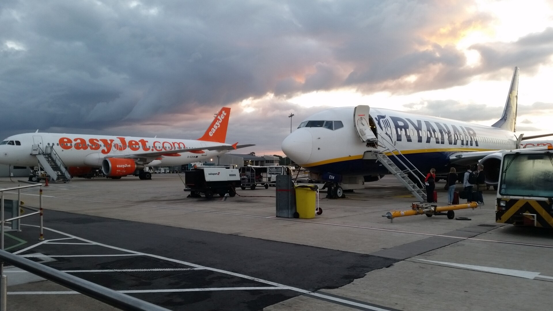 An easyJet and a Ryanair aircraft stand beside one another on the airport apron, preparing for people to board. The easyJet plane is on the left, the Ryanair plane on the right, both centrally pictured against a background of a cloudy sky with bright pink-yellow sunlight peeking through. The foreground shows the tarmac and concrete of the airport ground.