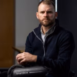Shane Hyhorec in a black jacket and a grey-stripped shirt underneath holding the SmartDrive wheelchair motor.