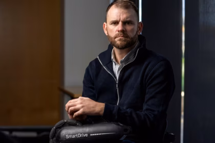 Shane Hyhorec in a black jacket and a grey-stripped shirt underneath holding the SmartDrive wheelchair motor.
