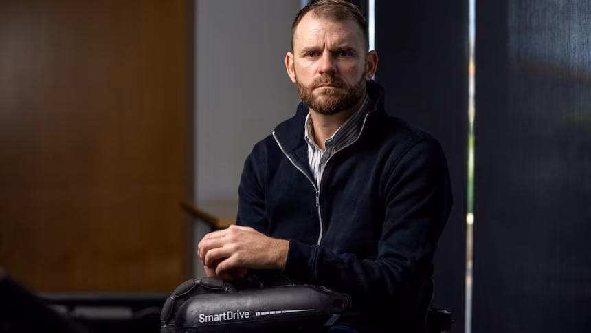 Shane Hyhorec in a black jacket and a grey-stripped shirt underneath holding the SmartDrive wheelchair motor.