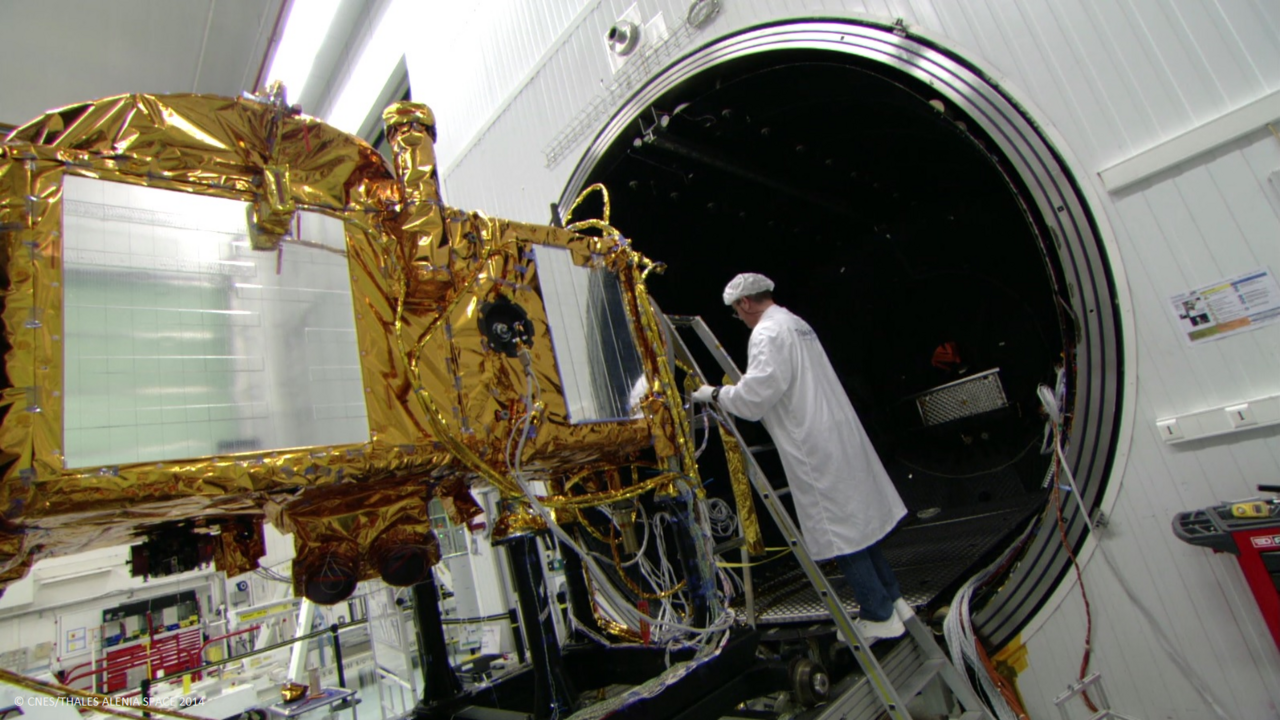 A gold and silver Thales Alenia satellite is moved through a large, round metal door into a cleanroom in Thales' France facility. A technician inspects from a ladder.