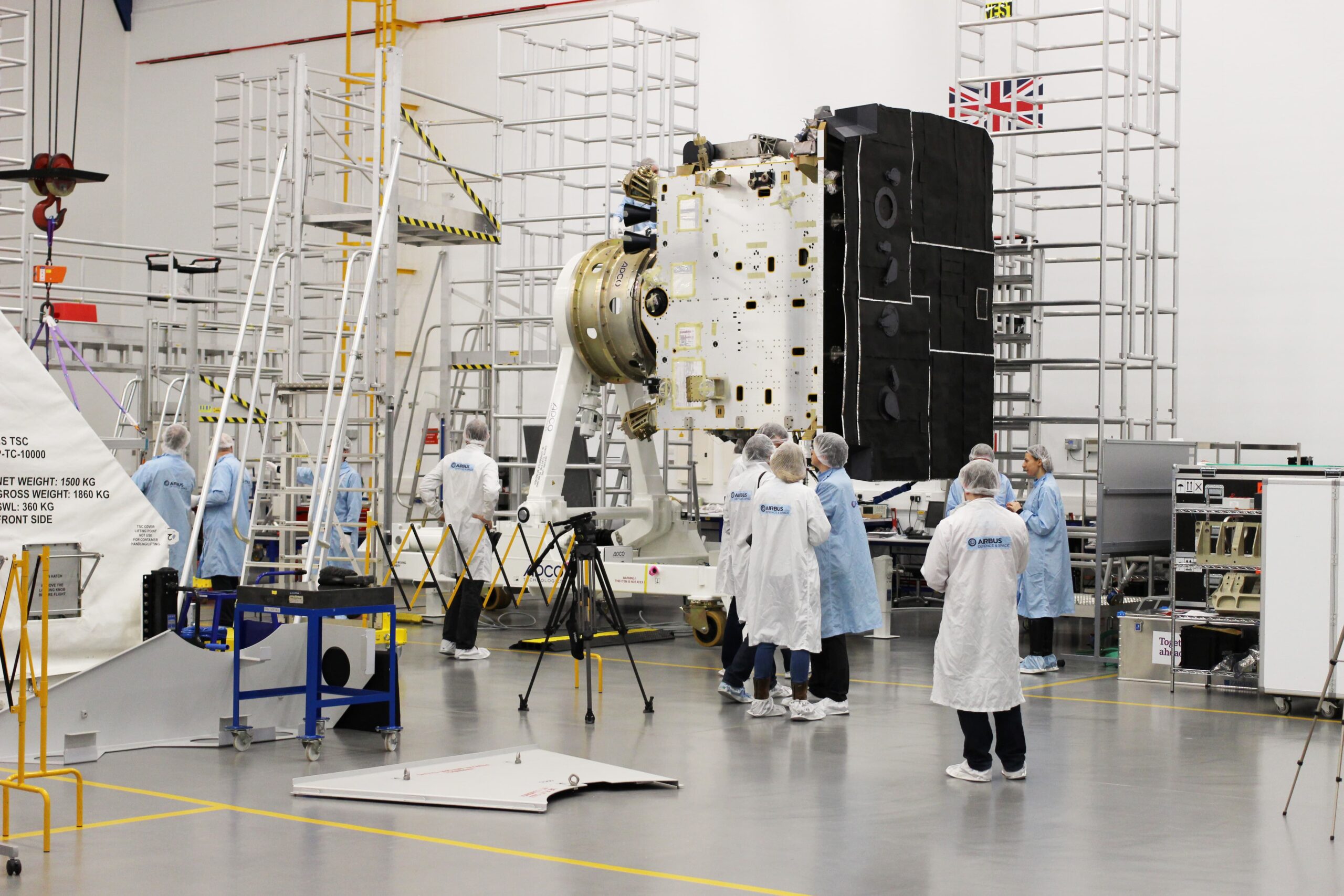 A large black and white cuboidal model of a solar orbiter mounted on scaffolding and being inspected by a group of people in a cleanroom at the Airbus Defence & Space Facility, Stevenage, UK.