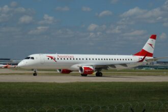 An aircraft rests on the runway. The backdrop is a blue sky with sparse clouds. Grass appears in the foreground.