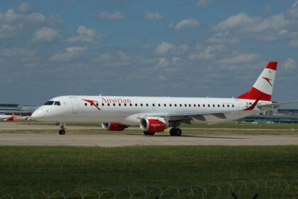 An aircraft rests on the runway. The backdrop is a blue sky with sparse clouds. Grass appears in the foreground.