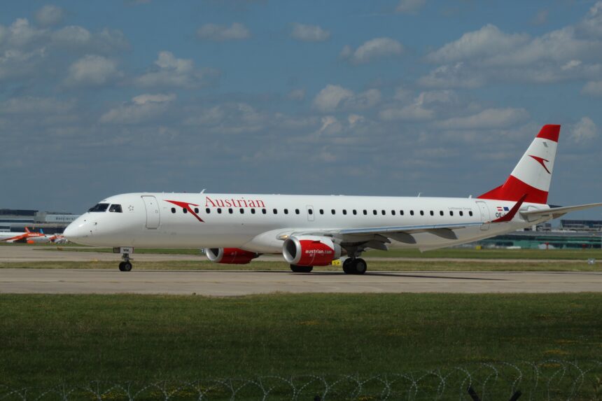 An aircraft rests on the runway. The backdrop is a blue sky with sparse clouds. Grass appears in the foreground.