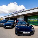 Exterior view of Norwich Airport terminal building with prominent “NorwichAirport” signage and the text “Part of Regional & City Airports” displayed above the entrance. Three airport transfer vehicles, including a Porsche, a Tesla and a Mercedes-Benz van branded for A2B Taxis Norwich, are parked outside the terminal. Additional signage for Europcar, Avis and Budget is visible on the building, with a bright blue sky and large white clouds overhead.