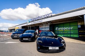 Exterior view of Norwich Airport terminal building with prominent “NorwichAirport” signage and the text “Part of Regional & City Airports” displayed above the entrance. Three airport transfer vehicles, including a Porsche, a Tesla and a Mercedes-Benz van branded for A2B Taxis Norwich, are parked outside the terminal. Additional signage for Europcar, Avis and Budget is visible on the building, with a bright blue sky and large white clouds overhead.