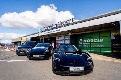 Exterior view of Norwich Airport terminal building with prominent “NorwichAirport” signage and the text “Part of Regional & City Airports” displayed above the entrance. Three airport transfer vehicles, including a Porsche, a Tesla and a Mercedes-Benz van branded for A2B Taxis Norwich, are parked outside the terminal. Additional signage for Europcar, Avis and Budget is visible on the building, with a bright blue sky and large white clouds overhead.