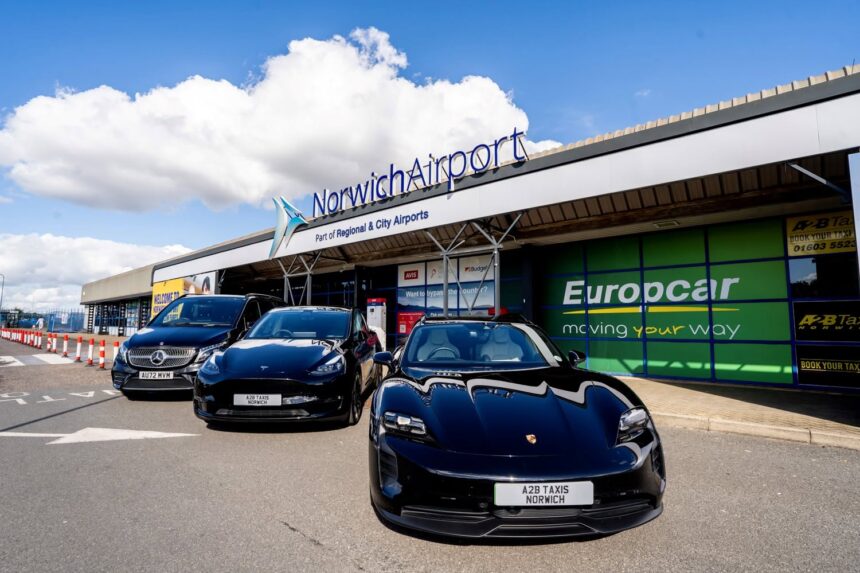 Exterior view of Norwich Airport terminal building with prominent “NorwichAirport” signage and the text “Part of Regional & City Airports” displayed above the entrance. Three airport transfer vehicles, including a Porsche, a Tesla and a Mercedes-Benz van branded for A2B Taxis Norwich, are parked outside the terminal. Additional signage for Europcar, Avis and Budget is visible on the building, with a bright blue sky and large white clouds overhead.