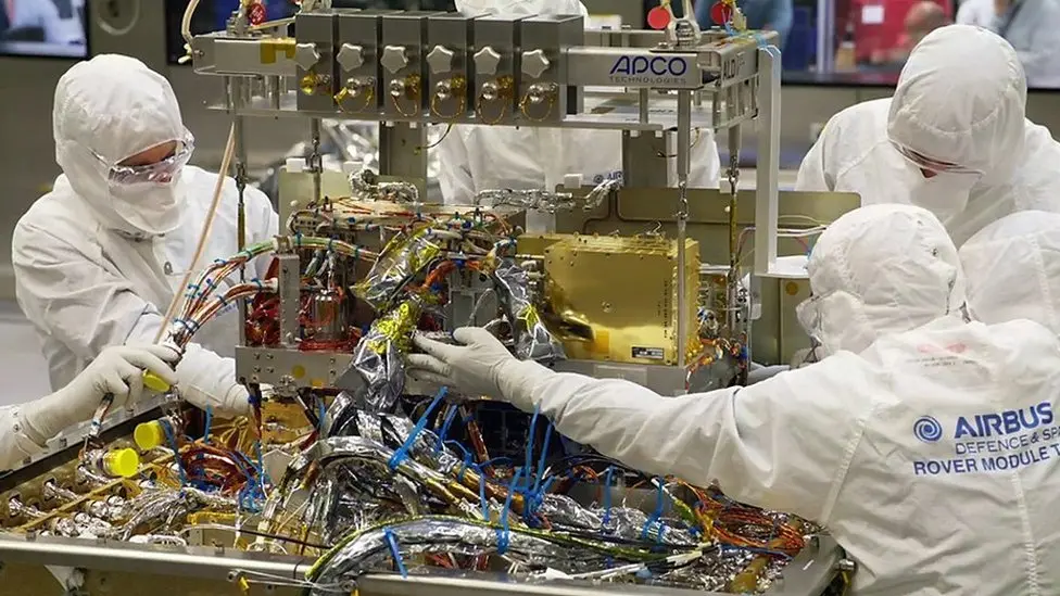 Five members of the Airbus team wearing coveralls, masks and goggles constructing some electrical components of Rosalind Franklin in their cleanroom.