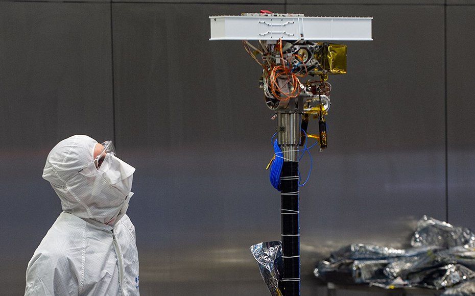 A technician wearing coveralls, a mask and goggles inspects the PanCam instrument – a long white box on the end of a black mast – mounted on the rover.