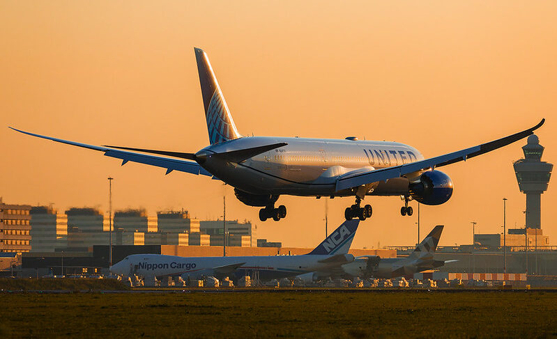 United Airlines aircraft landing at an airport at sunset, with the control tower and other aircraft visible in the background