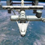Artist’s impression of the Dream Chaser spaceplane, which has a futuristic, streamlined, bulbous aircraft shape, docked to the ISS, a large satellite with many gantries and solar panels, orbiting above the ocean on Earth.