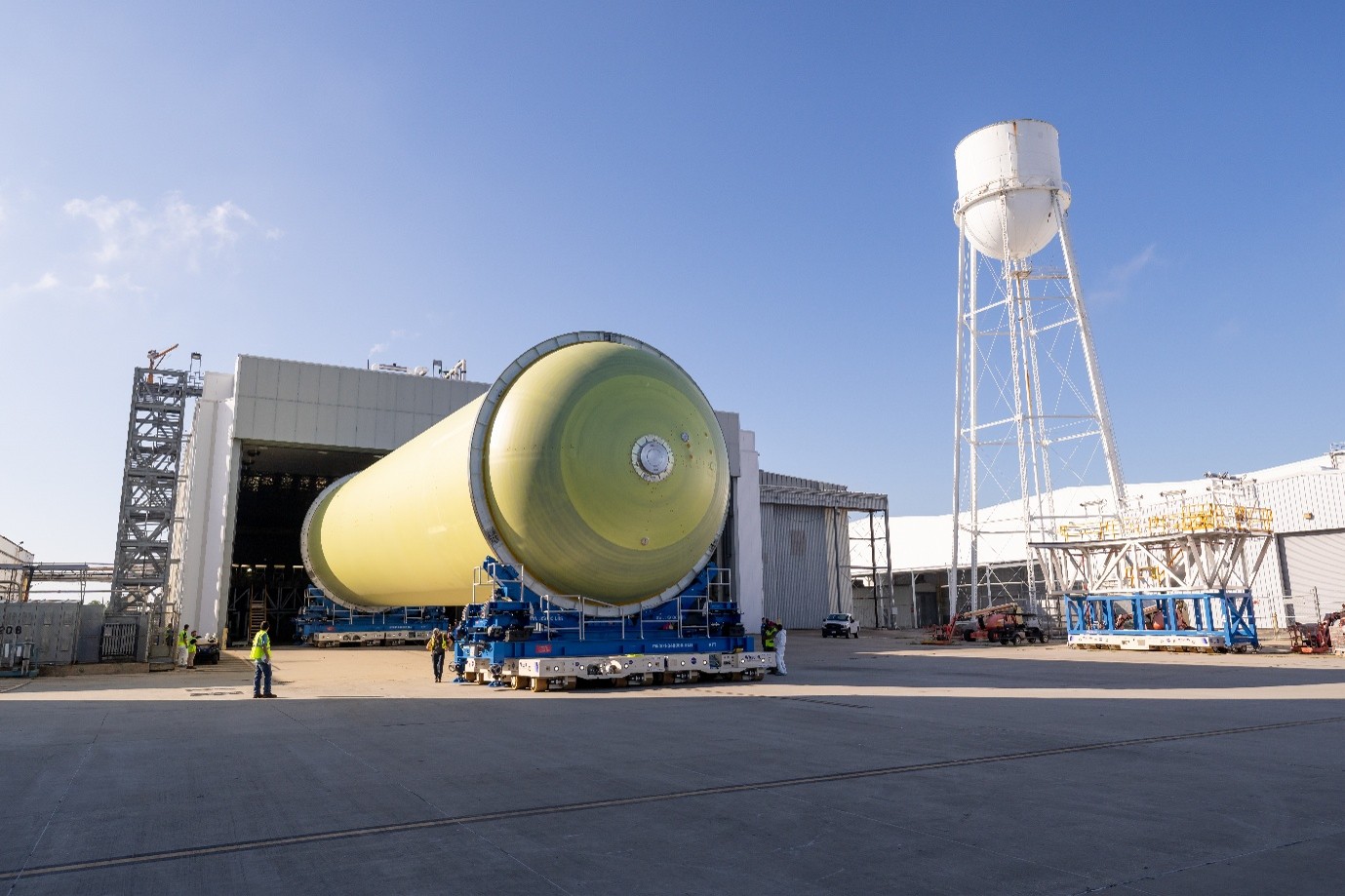 The huge grey cylindrical external tank for NASA’s Artemis III mission exits a large storage building before application of orange insulation to keep its liquid hydrogen contents cool. Staff members watch on and there is a large silo on stilts in the background.