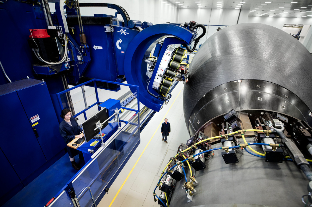 The robotic arm of a blue automated fibre placement machine, under the control of a technician, constructs the grey rounded composite fuselage for a Boeing 787 inside a white factory room