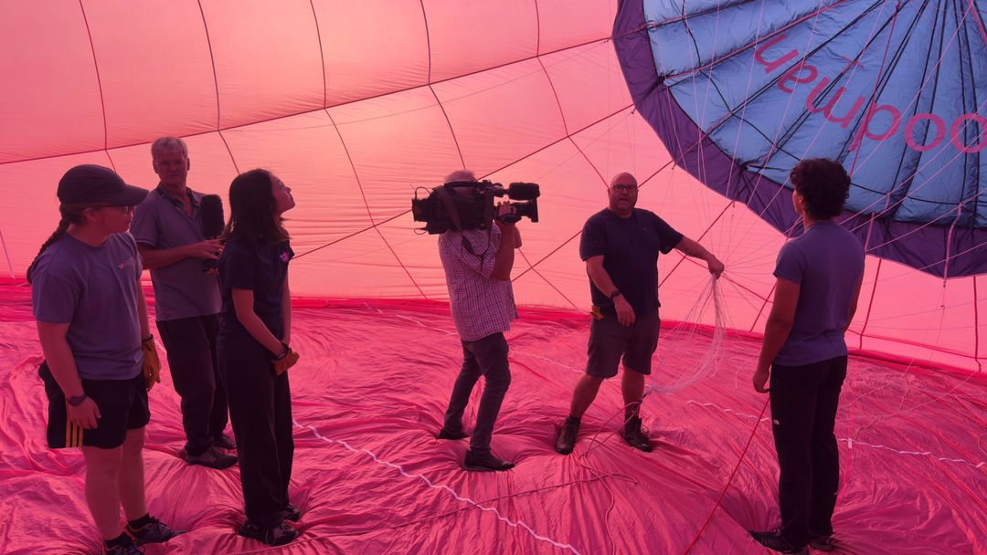 Image shows a number of people inside a red and blue hot air balloon with the pilot holding strings and being filmed by a video camera
