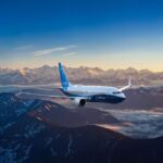 A Boeing 737 MAX with the Boeing label flying over a mountainous landscape, against a sunny horizon.
