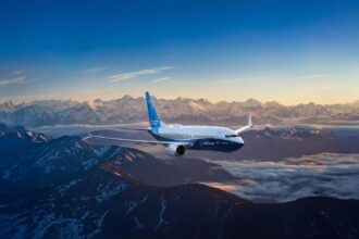 A Boeing 737 MAX with the Boeing label flying over a mountainous landscape, against a sunny horizon.