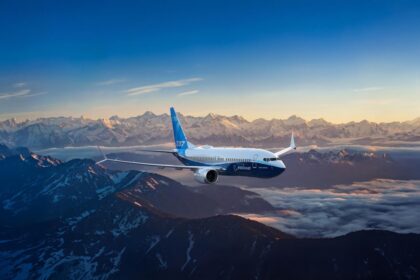 A Boeing 737 MAX with the Boeing label flying over a mountainous landscape, against a sunny horizon.