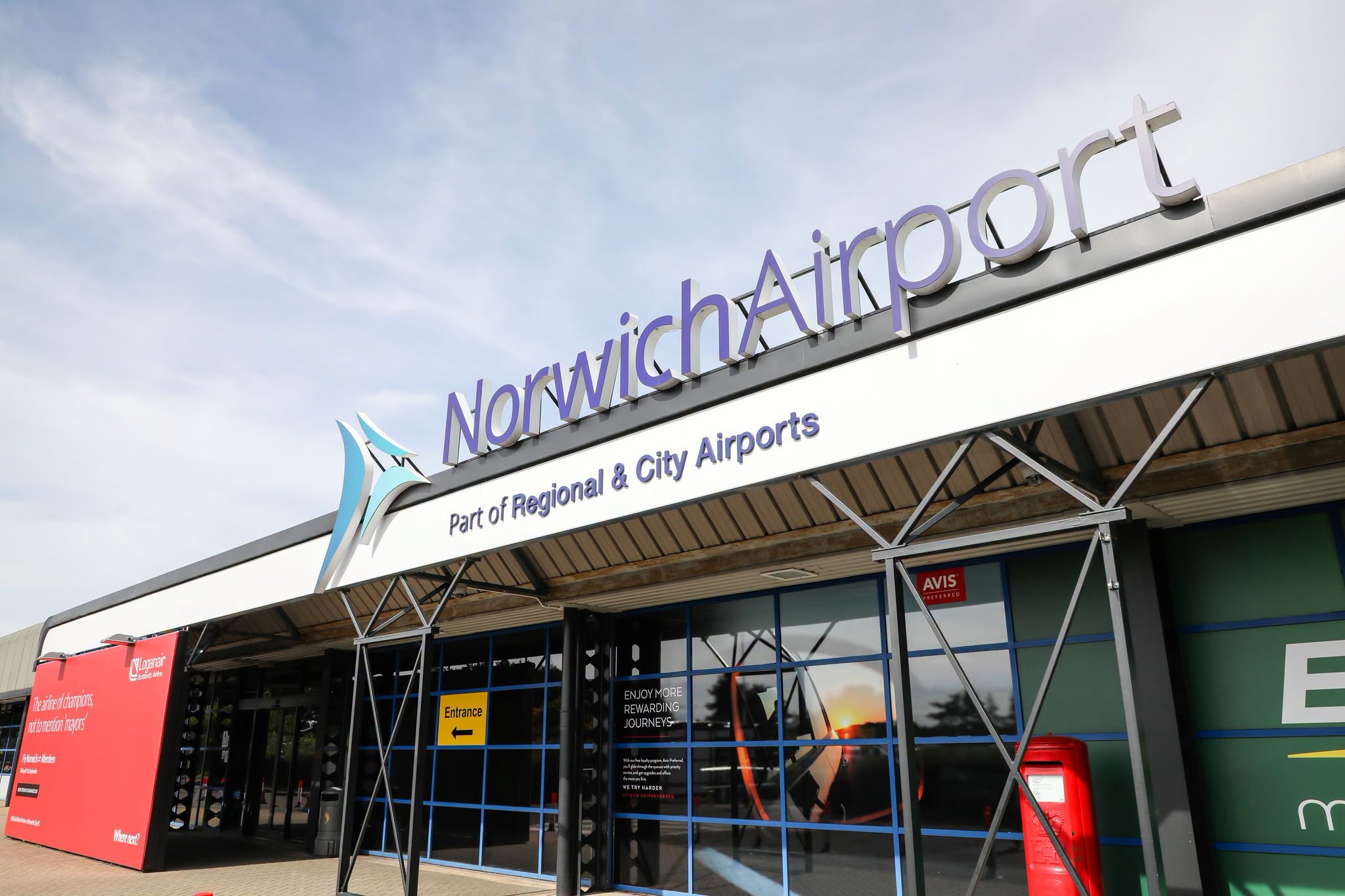 Exterior view of Norwich Airport terminal building, featuring large purple “NorwichAirport” signage above the entrance and the text “Part of Regional & City Airports” displayed on the facade. The building has blue-framed glass windows, steel support structures, and visible airport service signage, including an entrance sign and an Avis rental counter sign. The scene is photographed in daylight under a lightly clouded sky