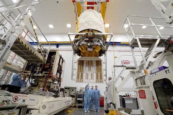 The Aeolus satellite suspended from the ceiling and shrouded in white material while being assembled in a cleanroom at Airbus’ Stevenage facility. Several technicians in protective clothing look on, and are surrounded by machinery and electronics.