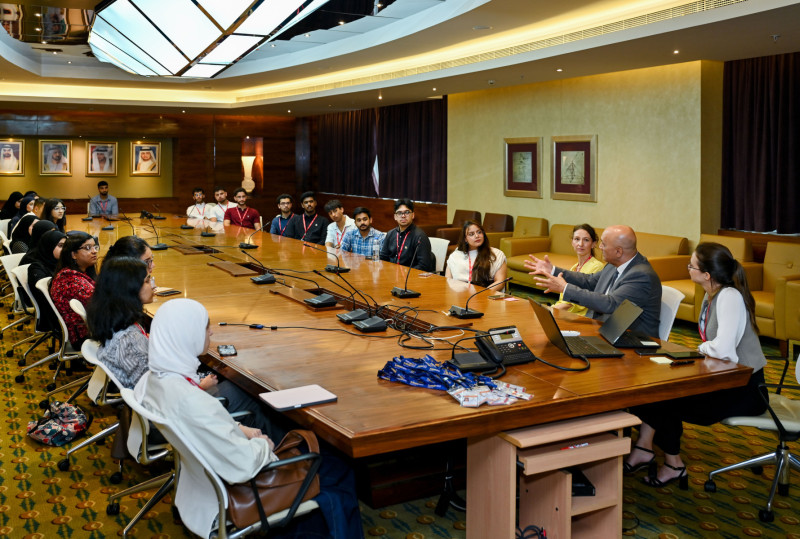 Students sit around a large wooden conference table listening to a man. the table has microphones and is in a large brown coloured room with paintings on the walls.