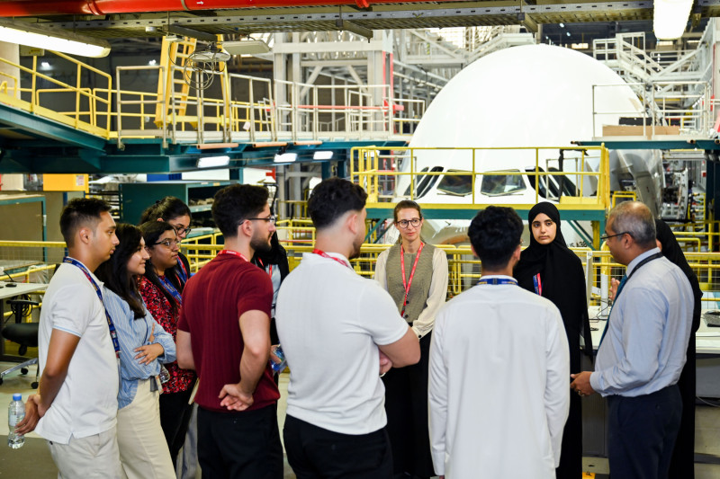 Students stand in a circle in a factory type facility with machinery in the background, listening to a male emirates representative.