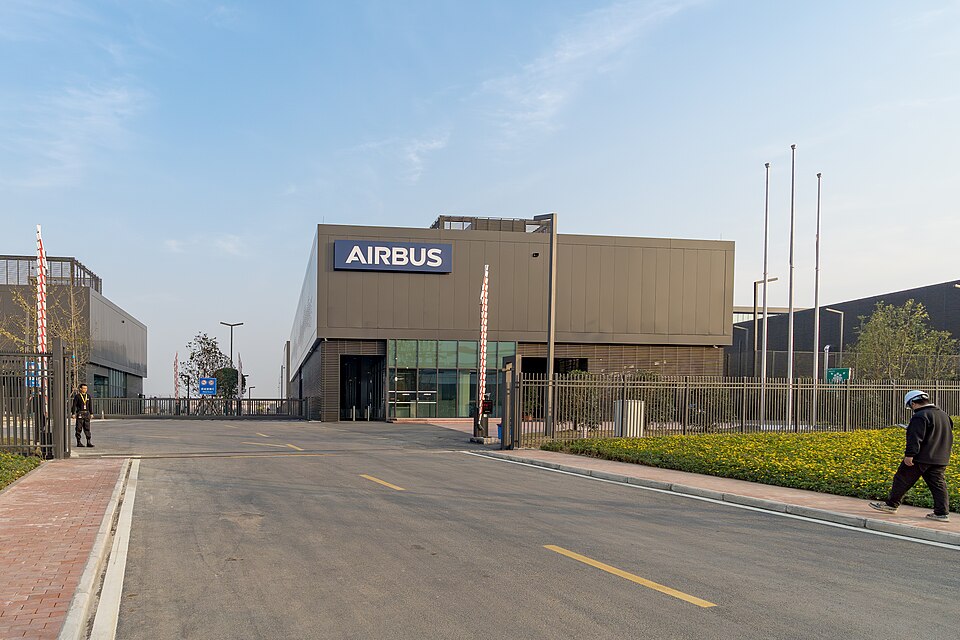 An exterior view of a modern Airbus facility featuring a large, boxy, dark grey industrial building with a prominent blue "AIRBUS" sign on its upper facade. An asphalt road leads toward a gated entrance with raised red-and-white barriers. A security guard stands to the left of the gate, while on the right, a person wearing a dark jacket and a white hard hat walks along a sidewalk next to a landscaped bed of yellow flowers under a clear blue sky.