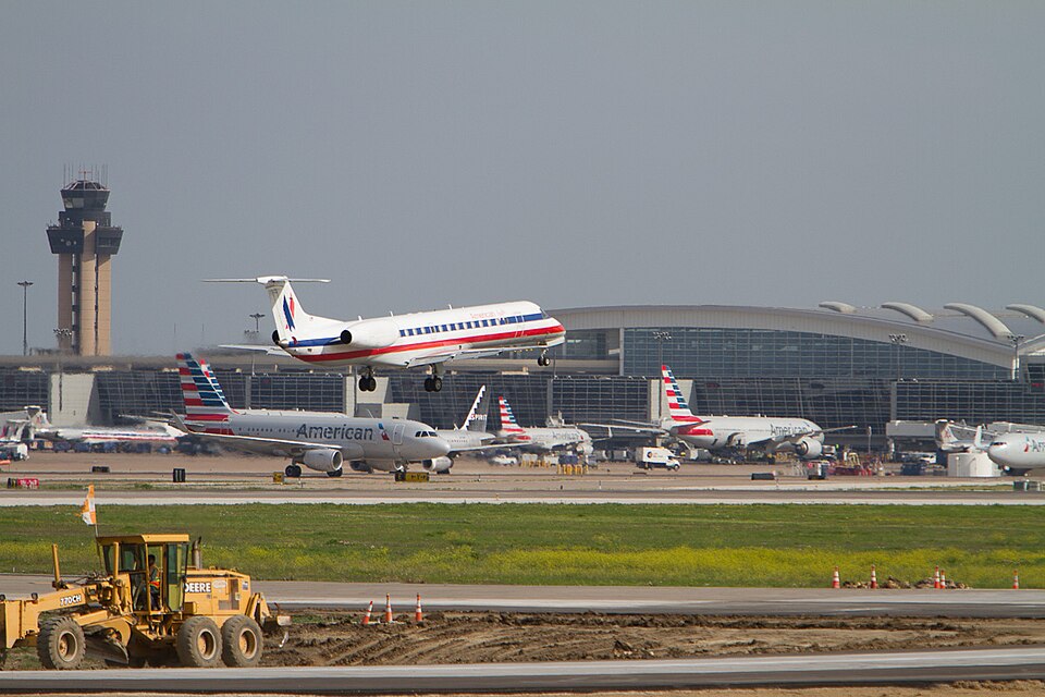 Both American Airlines and American Eagles aircraft parked and departing outside of Dallas Fort Worth International Airport. The sky is grey and the viewer may see the control tower and a terminal.