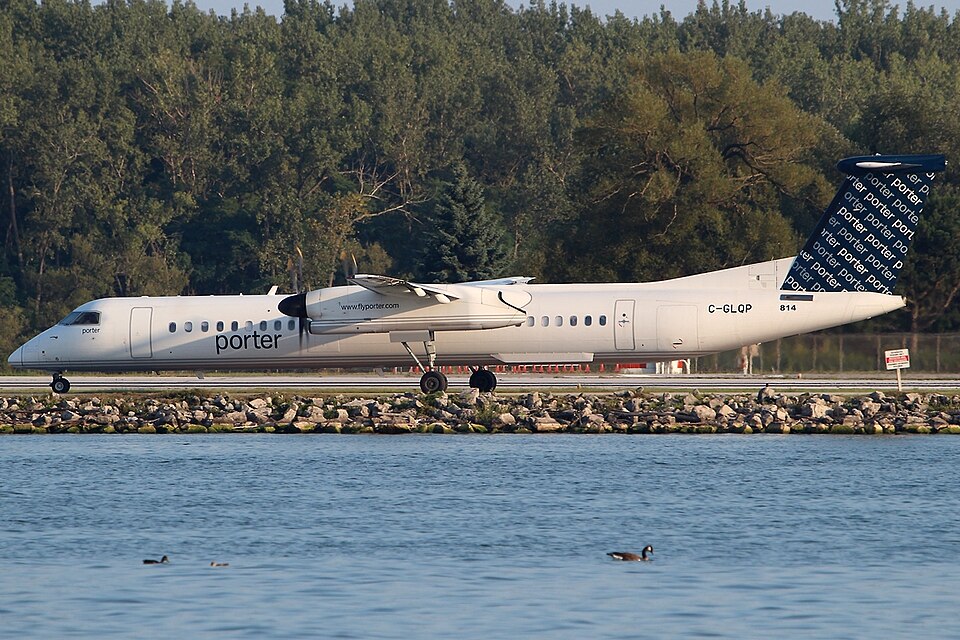 A Porter Airlines aircraft after landing. The aircraft is behind a lake and in front of several pine trees, with the blue sky peaking through the tops of the trees. The livery is mostly white.