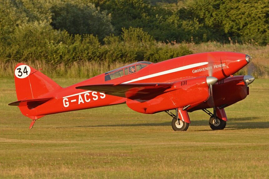 Image shows a grounded red De Havilland DH88 Comet ‘G-ACSS’ aircraft during the day