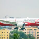 A Qantas Boeing 787-9 Dreamliner (registration VH-ZNH) in the special "Qantas 100" centenary livery on final approach with landing gear extended, set against a backdrop of dense urban buildings.