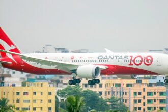 A Qantas Boeing 787-9 Dreamliner (registration VH-ZNH) in the special "Qantas 100" centenary livery on final approach with landing gear extended, set against a backdrop of dense urban buildings.