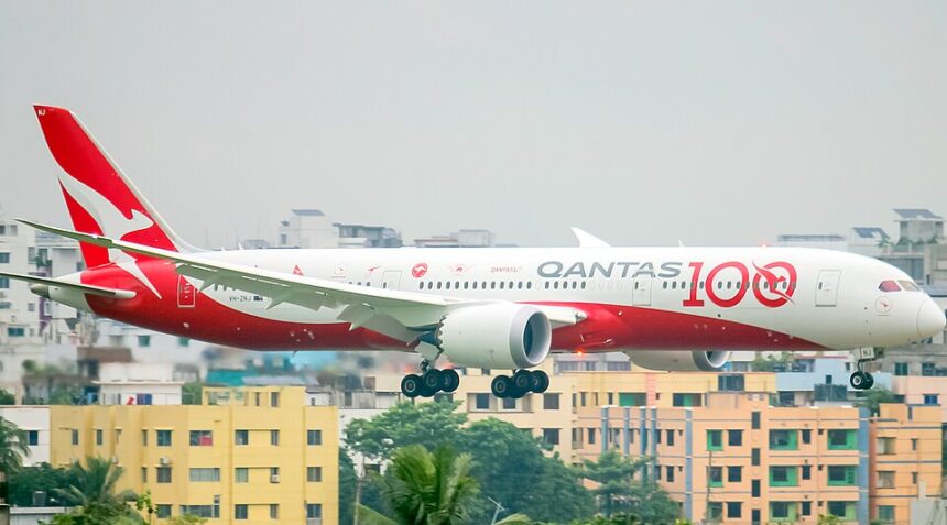 A Qantas Boeing 787-9 Dreamliner (registration VH-ZNH) in the special "Qantas 100" centenary livery on final approach with landing gear extended, set against a backdrop of dense urban buildings.