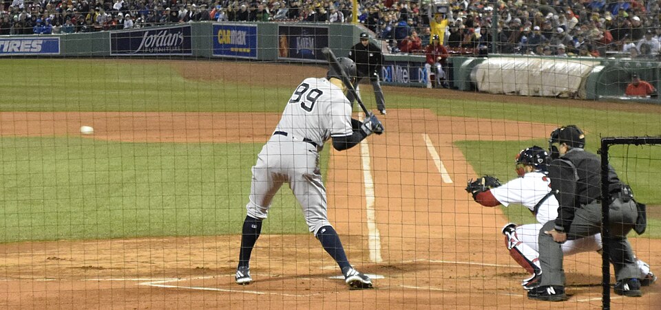Viewed through the protective netting behind home plate, a baseball player wearing a grey New York Yankees away uniform with the number 99 is in a batting stance, awaiting a pitch. A white baseball is visible in mid-air over the infield grass, approaching the plate. Behind the batter, a catcher in a white and red home uniform crouches in position, with a black-clad umpire stationed directly behind him. The scene is set in a crowded stadium at night, with advertising signs and spectators visible in the background.