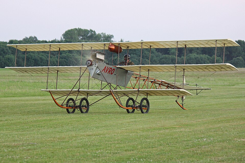 Image shows a replica Avro Triplane on the grass during the day at Shuttleworth in 2017 with pilot and moving propellers visible
